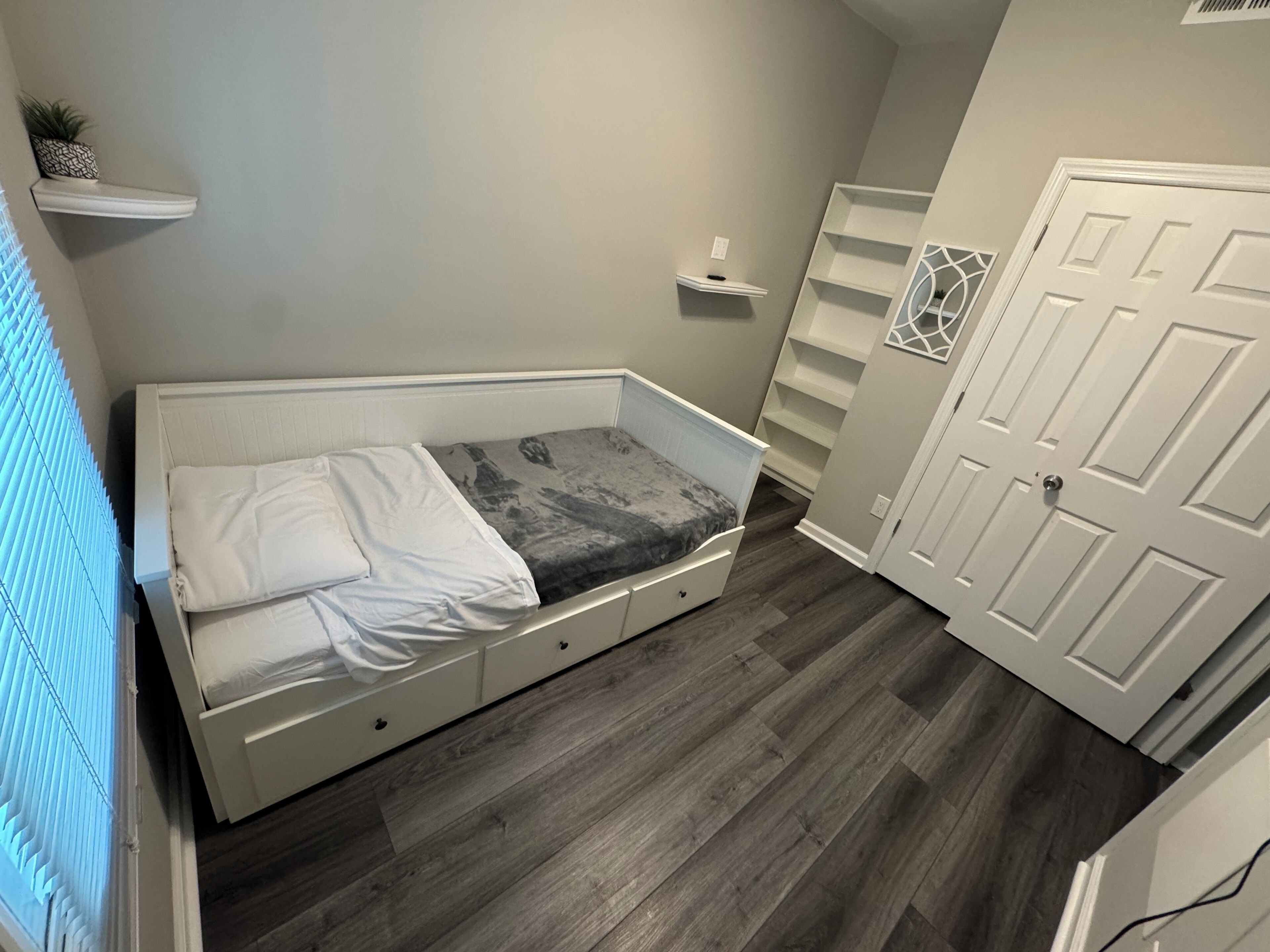 Ein Schlafzimmer mit einem Bett, einer kleinen Ablagefläche, einer Tür und einem Fenster mit Jalousien.