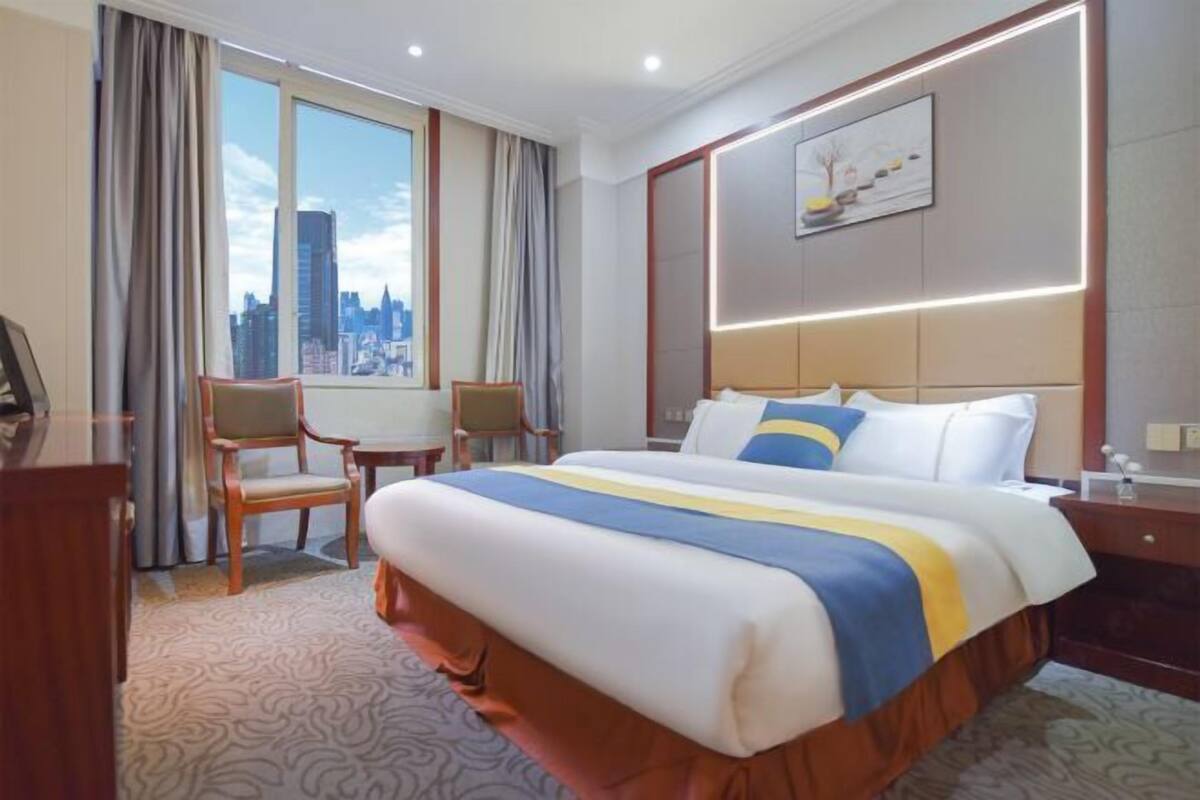 A hotel room with a large bed, a desk, and a chair. There is a window with a view of a city skyline.