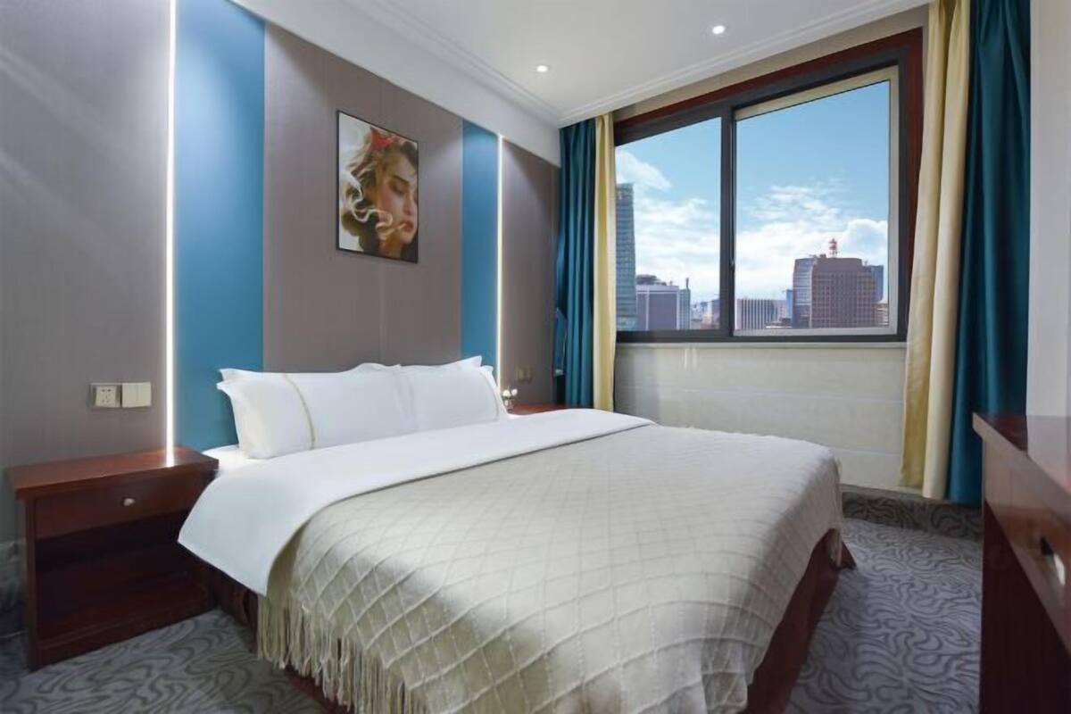 A hotel room with a large bed, a nightstand, a window with a city view, and a painting on the wall.