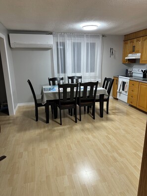 A dining area with a table and chairs, a kitchen with wooden cabinets, and a window with sheer curtains.