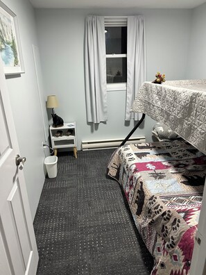 A small, compact room with a bunk bed, a small table with a lamp, a window with curtains, and a door.