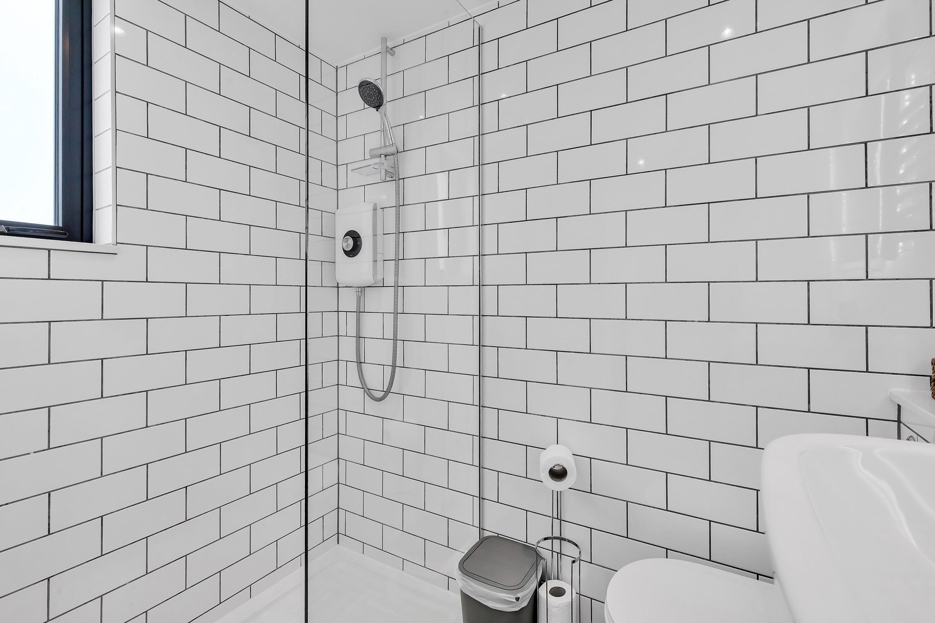 A bathroom with white subway tiles, a shower with a handheld showerhead, a toilet, and a trash can.