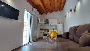 Un espacio habitable funcional con un sofá marrón, una zona de comedor con sillas amarillas, una cocina con armarios blancos y un televisor de pantalla plana montado en la pared.