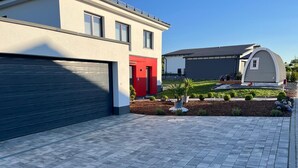 A modern residential area with a paved driveway leading to a garage, a red door, and a landscaped garden with potted plants.