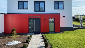 A modern two-story house with a red facade, large windows, and a small garden with a pathway.