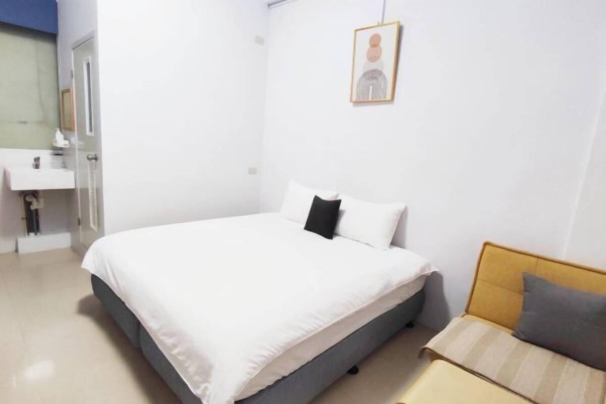 Un dormitorio con cama, mesita de noche, un pequeño sofá, un cuadro en la pared y un baño con lavabo y espejo.