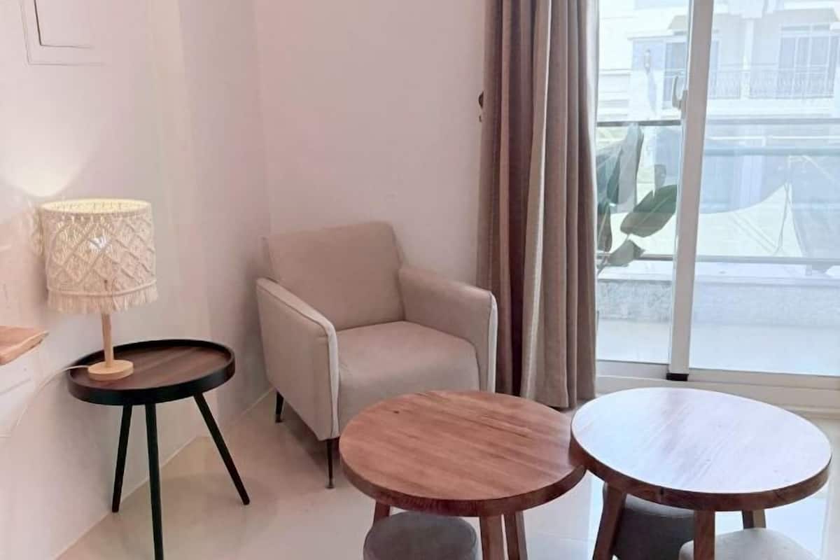 Una habitación con un sillón beige, dos mesas redondas de madera, una mesita auxiliar con una lámpara y un gran ventanal con cortinas.