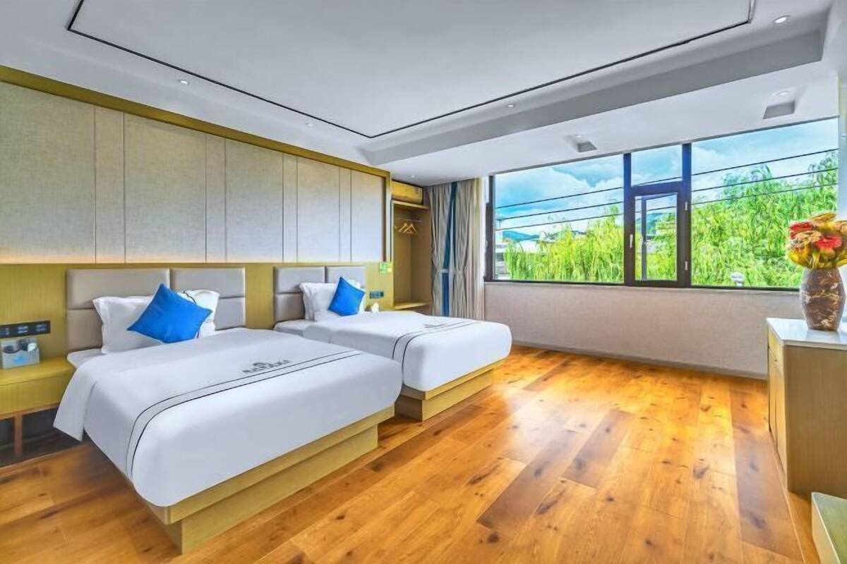 Hotelkamer met twee bedden, houten vloer, groot raam met uitzicht op groen en een bureau met vaas.