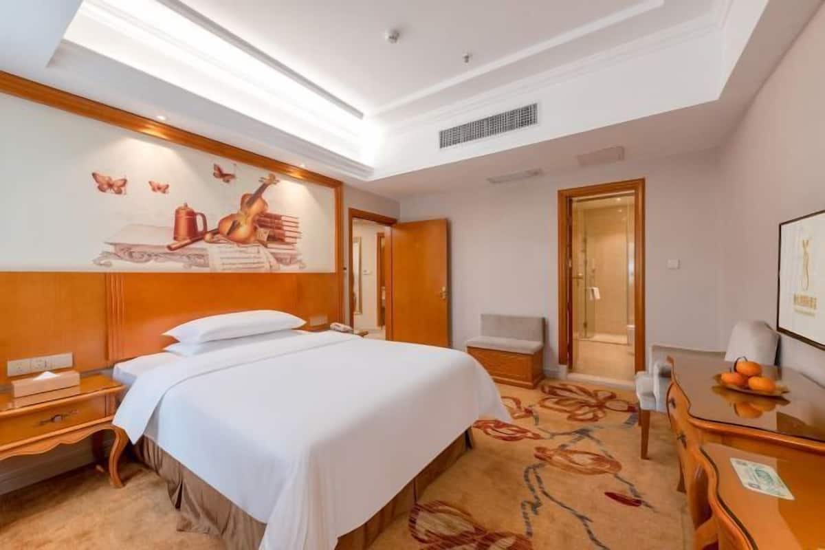 A hotel room with a large bed, a desk, a chair, a small table, and a wall-mounted TV.