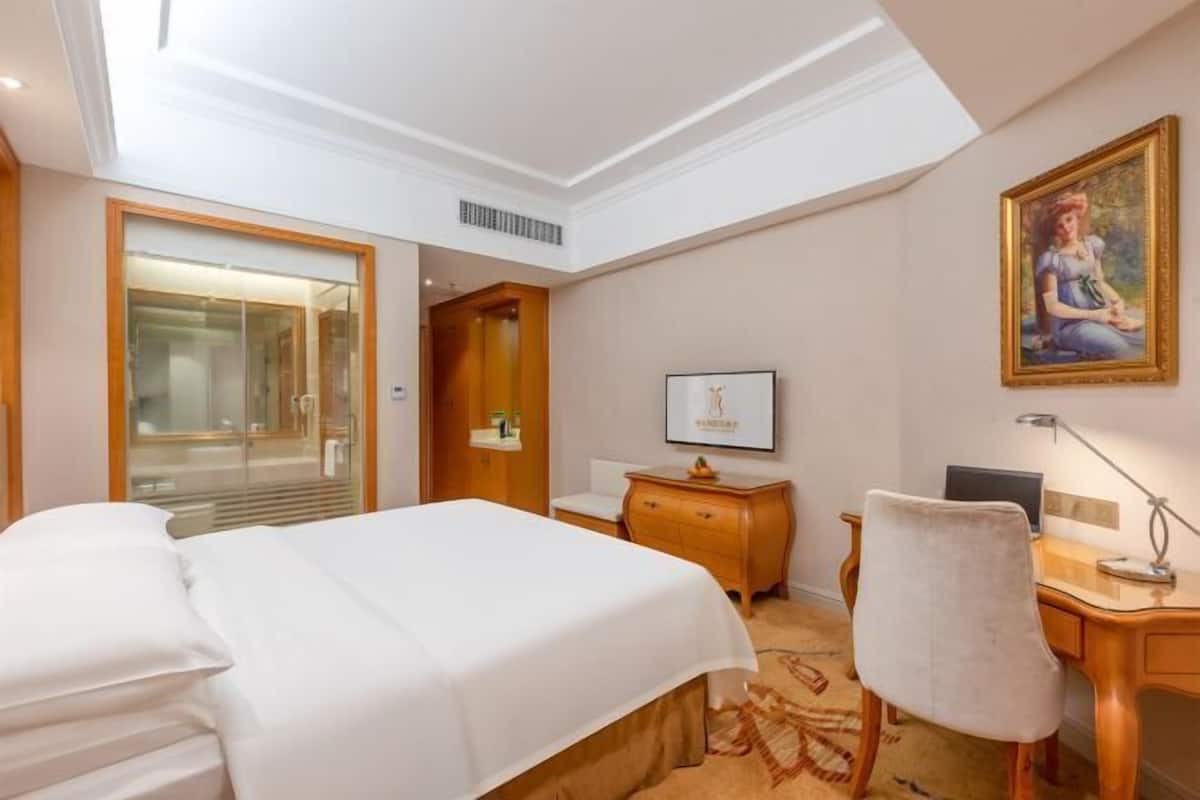 A hotel room with a bed, a desk, a chair, a painting on the wall, and a door leading to a bathroom.