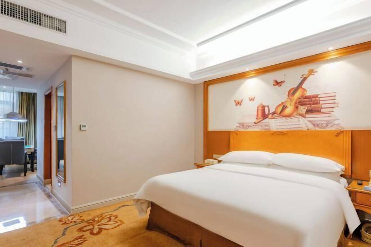 A hotel room with a bed, a nightstand, a wall with a guitar and books mural, a carpet, and a hallway leading to a dining area.