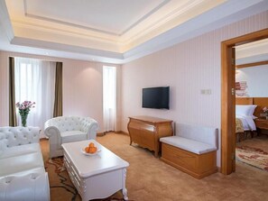 A hotel room with a white sofa, a white armchair, a wooden side table, a flat-screen TV mounted on the wall, and a wooden bed frame. - Vienna International Hotel (Guangzhou)