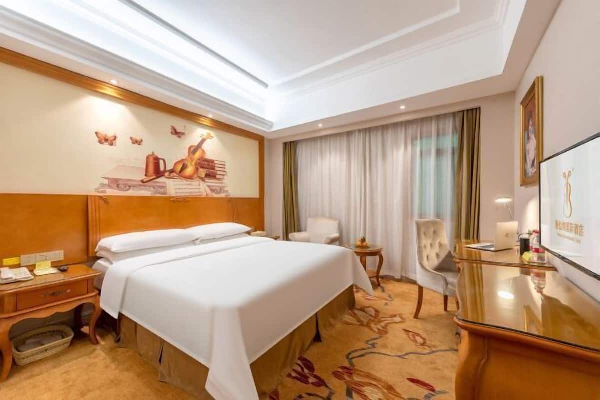 A hotel room with a large bed, a desk with a chair, a small table, and a television.