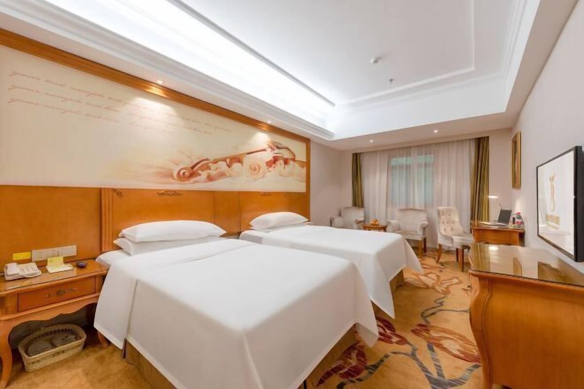 A hotel room with two beds, a desk, a chair, a desk lamp, and a large wall mural.