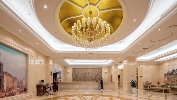 A spacious lobby with a large chandelier, a reception desk, and a mural on the wall. - Vienna International Hotel (Guangzhou Xintang High Speed Railway Station Cowboy City Branch) (Guangzhou)