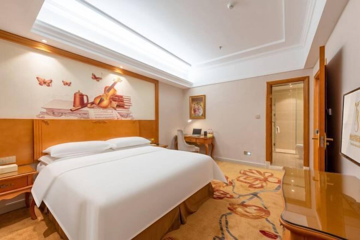 A hotel room with a bed, a desk, a chair, a small table, a painting, and a door.