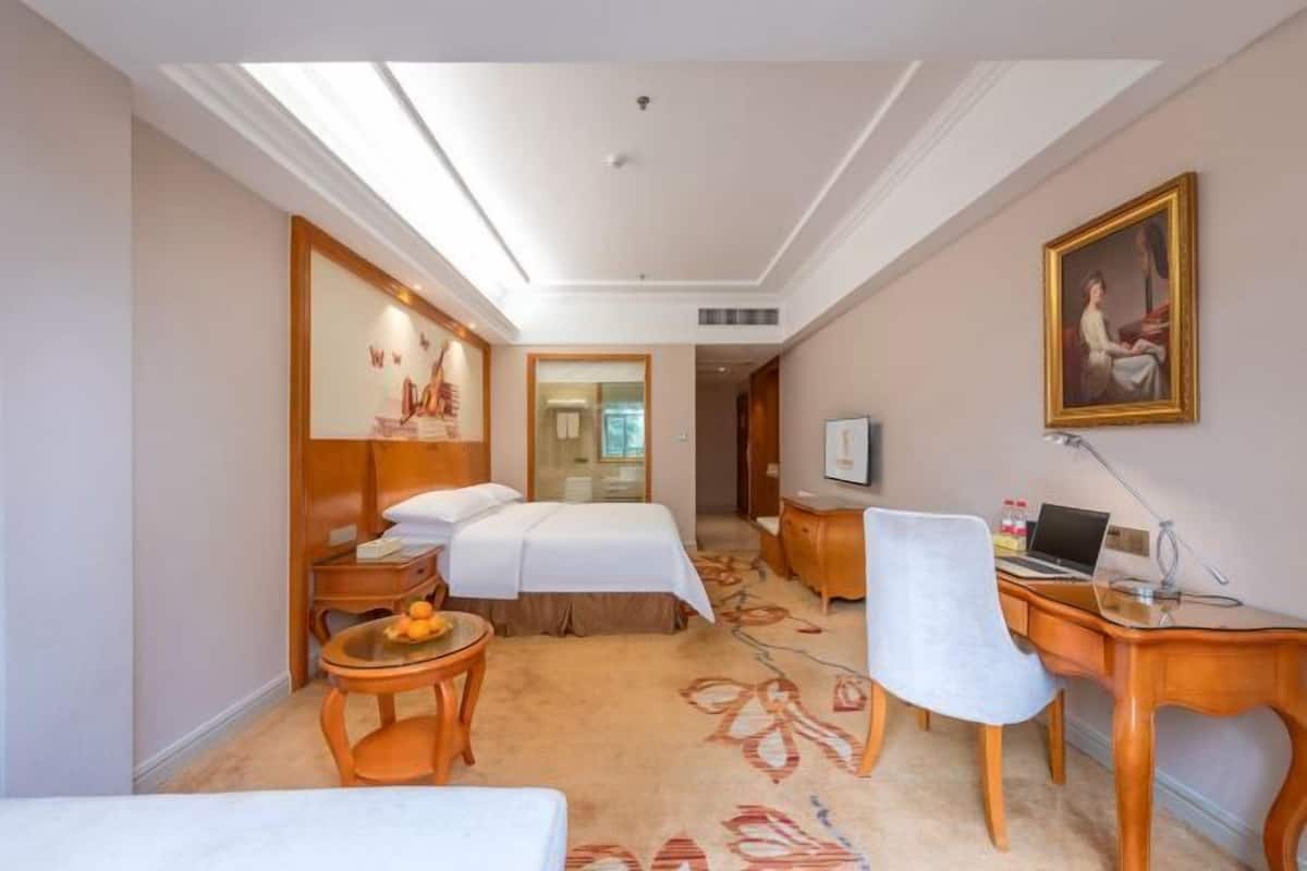 A hotel room with two beds, a desk, a chair, a small table, and a painting on the wall.