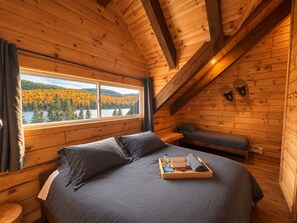 Une pièce de cabane en bois comprenant un lit, un banc et une fenêtre donnant sur un lac bordé de forêts.