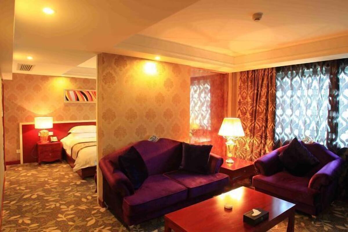 A hotel room with a purple sofa set, a wooden coffee table, a bed with a red headboard, and patterned curtains.