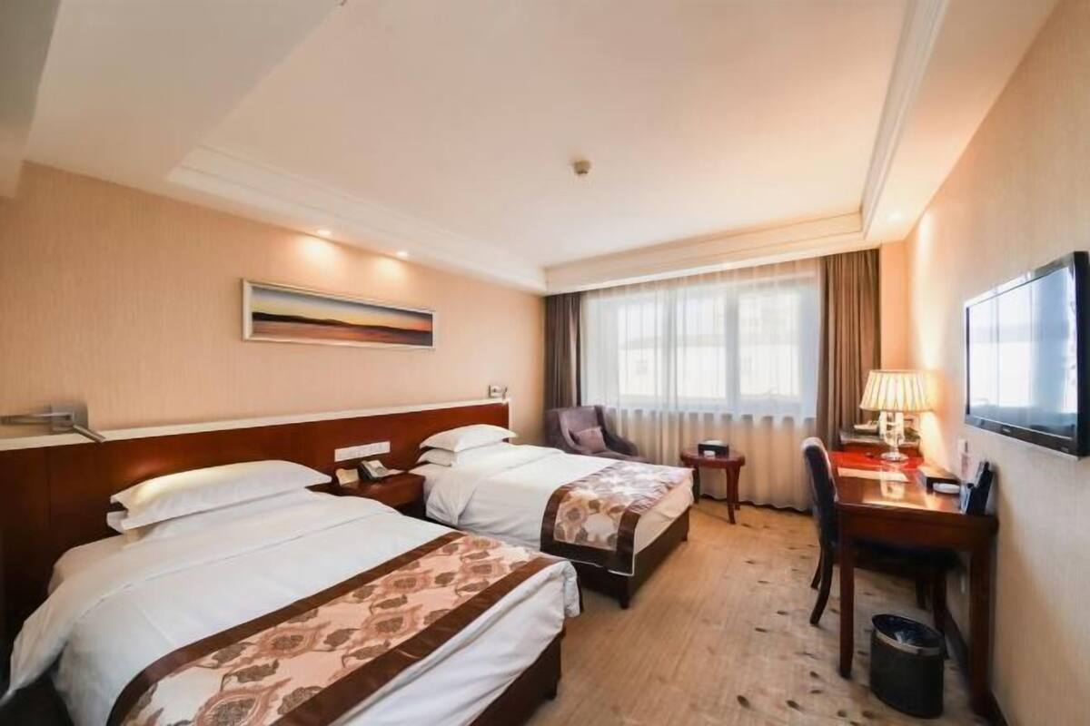 A hotel room with two beds, a desk, a chair, a lamp, and a television.