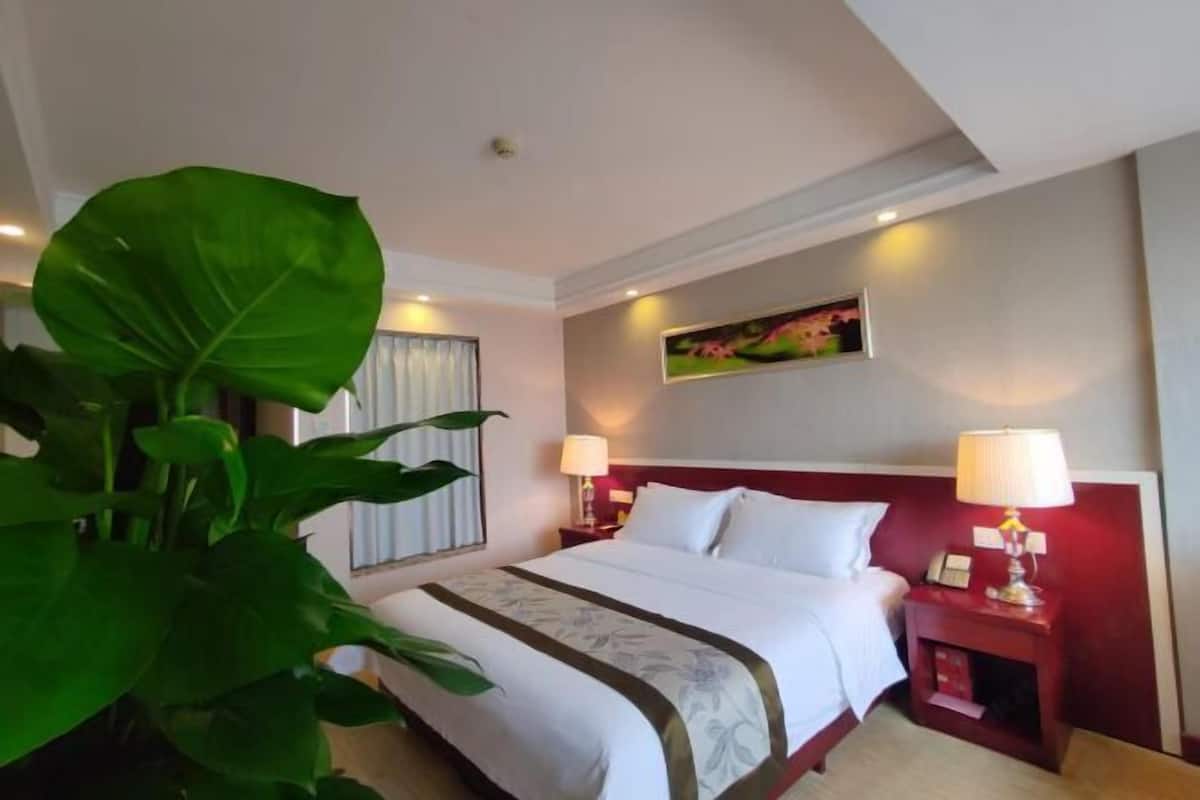 A hotel room with a large bed, two bedside tables with lamps, a painting on the wall, and a large green plant.