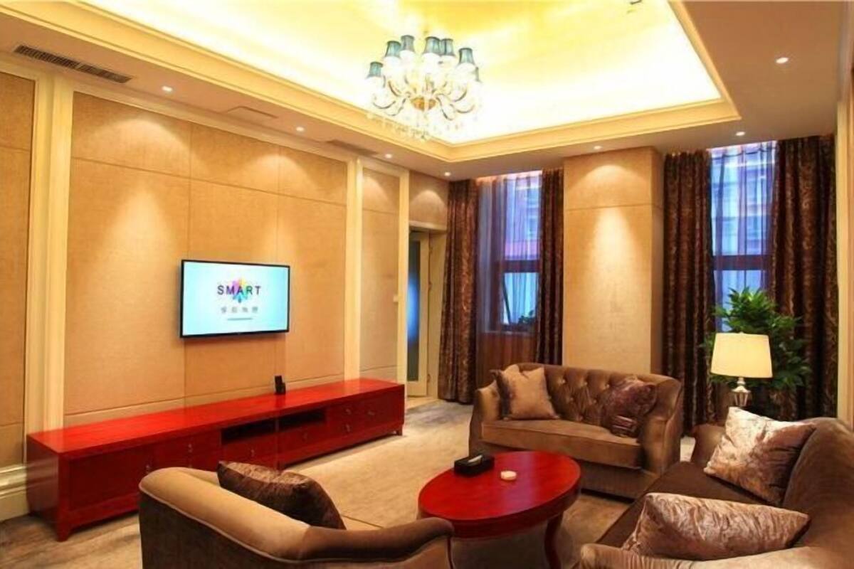 A hotel living room with a flat-screen TV, a red entertainment unit, leather sofas, a red coffee table, and a chandelier.