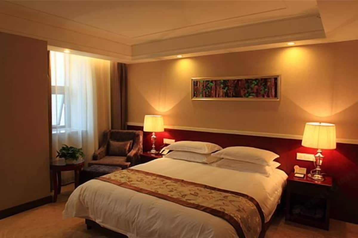 A hotel room with a large bed, two bedside tables with lamps, a chair, a painting on the wall, and a window with curtains.