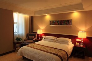 Habitación de hotel con una cama grande, dos mesitas de noche con lámparas, una silla, un cuadro en la pared y una ventana con cortinas.
