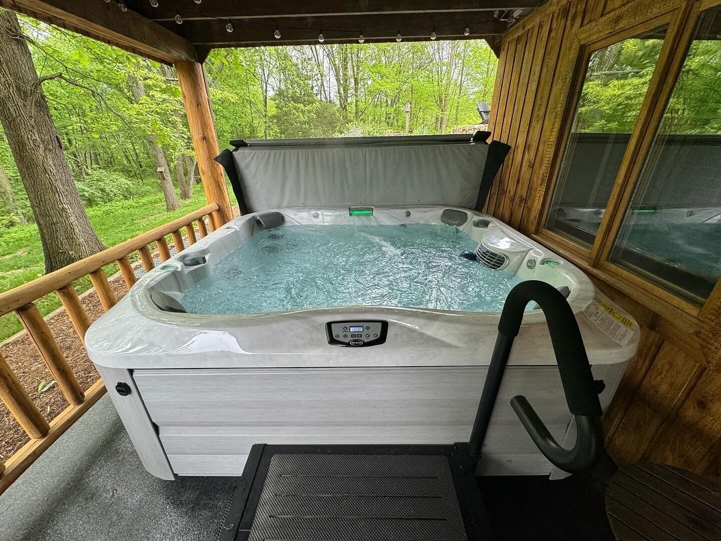 A hot tub with a control panel, surrounded by a wooden deck and a glass enclosure, overlooking a forested area.