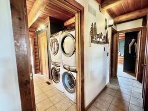 A laundry room with stacked washers and dryers, wooden beams, and a decorative tree wall art. - Serene Private Luxury Escape w/Beautiful Porches, Spa Patio & Fun Game Rooms (Bloomington)