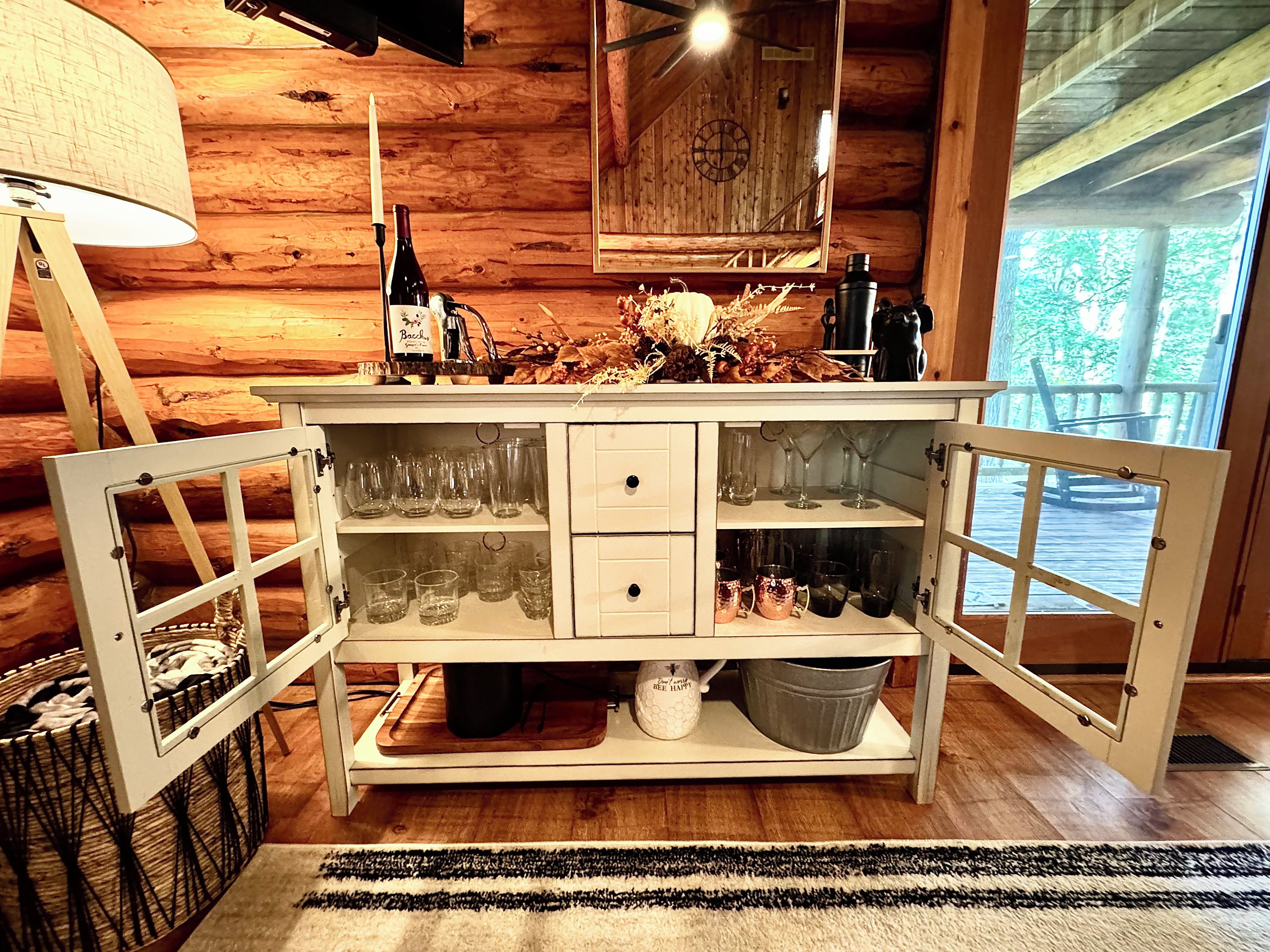 A wooden cabin interior with a white cabinet containing glasses and bottles, a lamp, and a view of the outdoors.