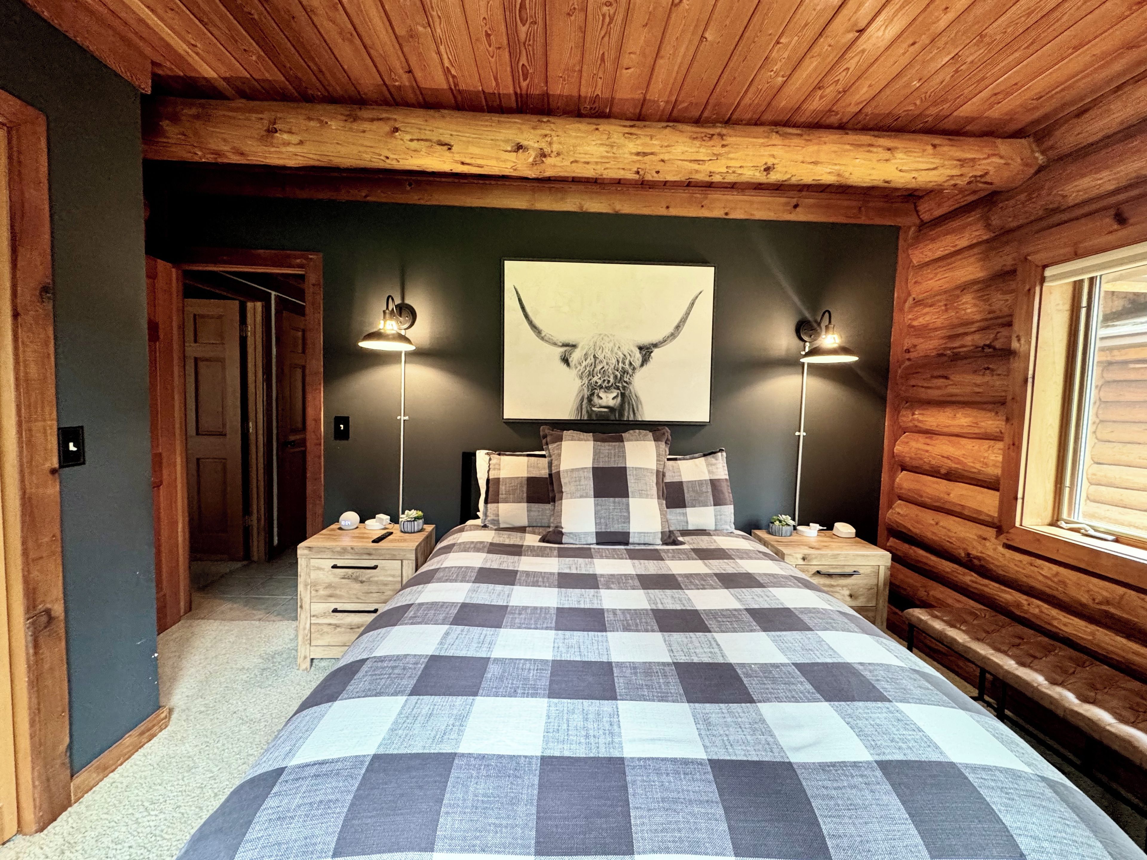 A bedroom with a checkered bedspread, two bedside lamps, a mounted artwork, and a wooden ceiling.