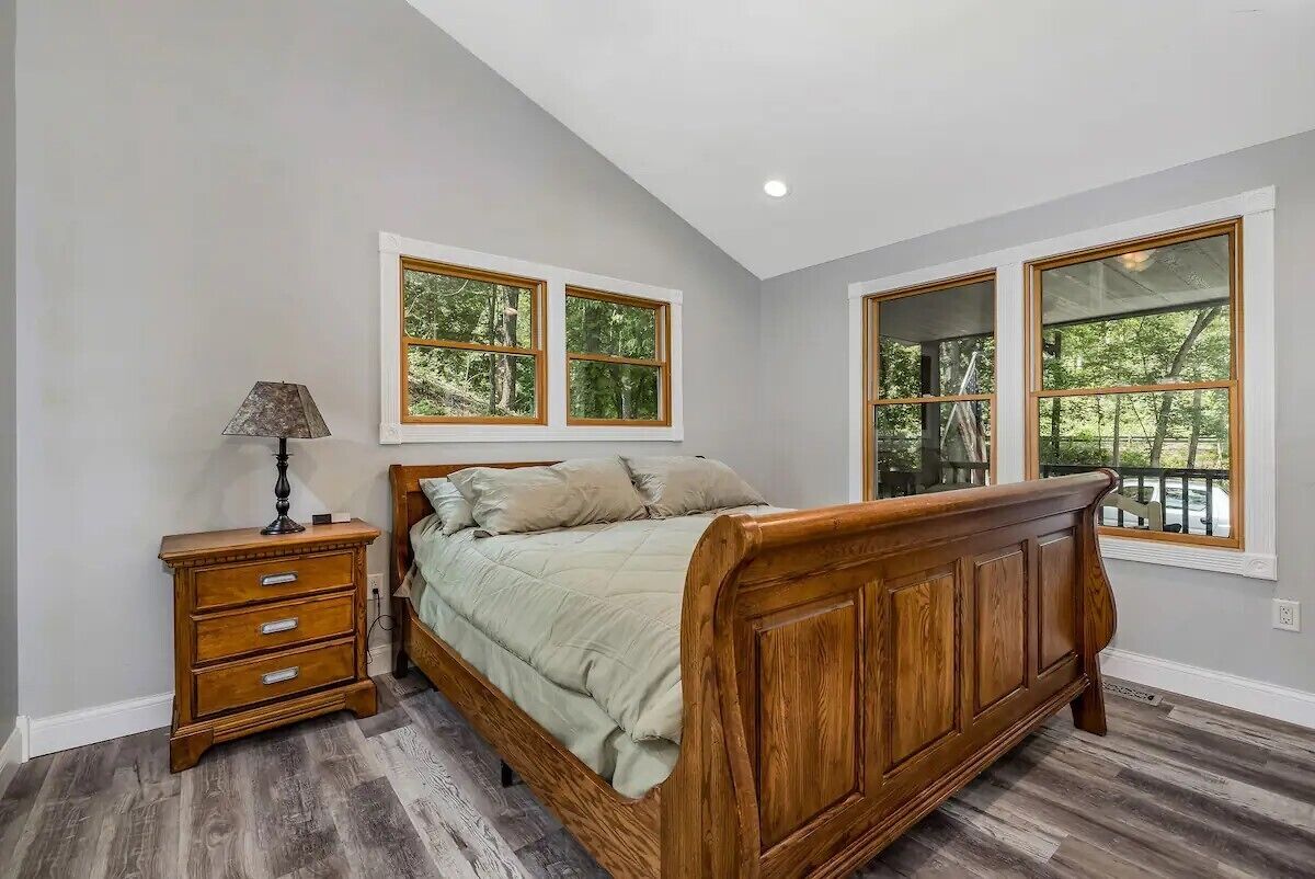 A bedroom with a wooden bed, a nightstand, and two windows with a view of trees.