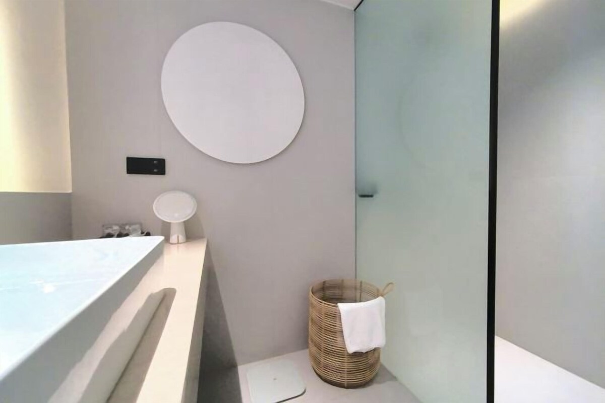 A bathroom with a bathtub, a round mirror, a wall-mounted light, and a wicker basket with a towel.