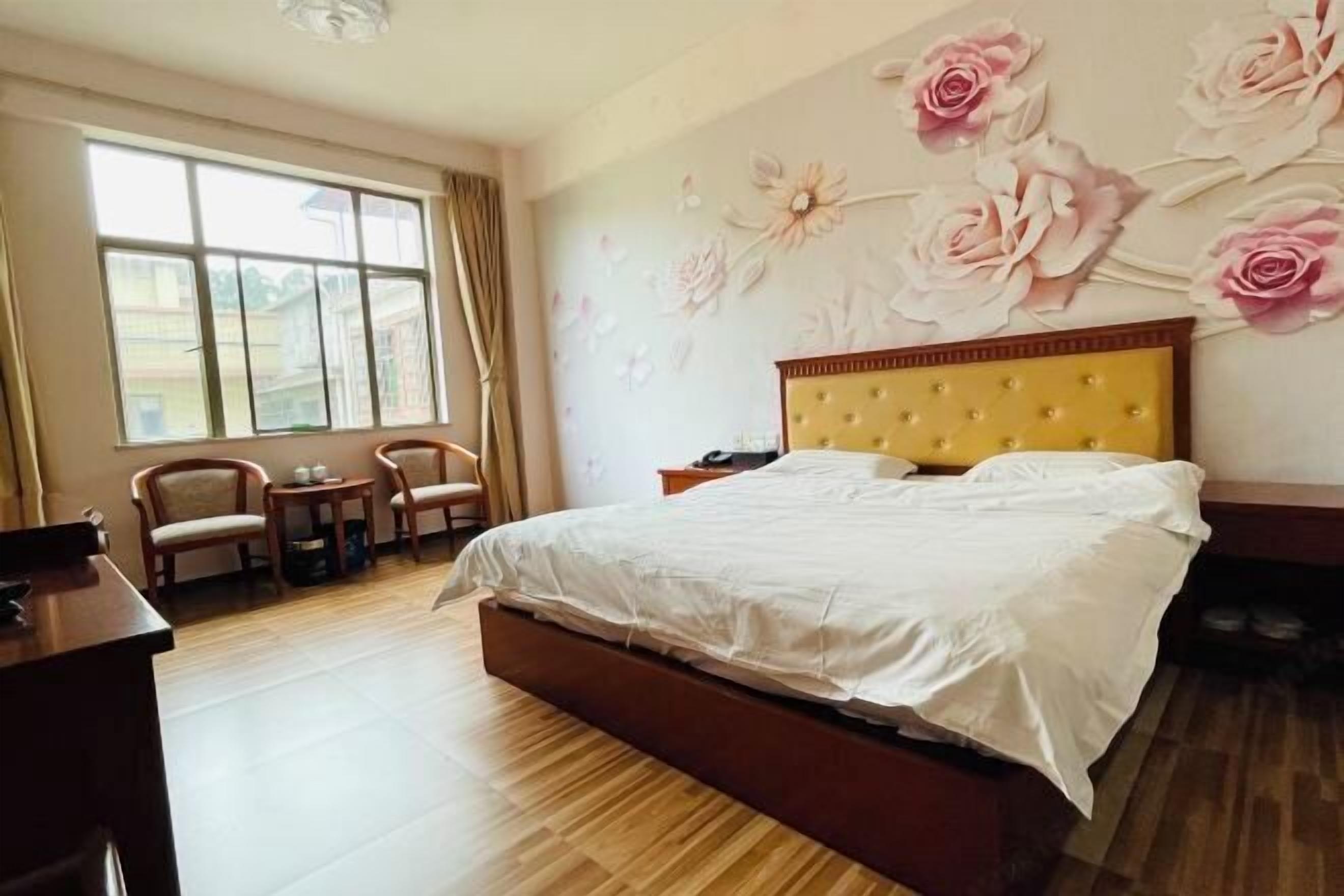 A bedroom with a bed, a desk, two chairs, a window, and floral wall art.