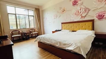 A bedroom with a bed, a desk, two chairs, a window, and floral wall art.