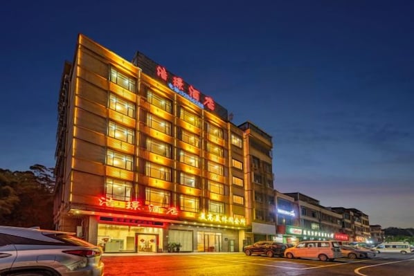 En hotelbygning med flere etager, oplyst skiltning, en parkeringsplads og flere biler.