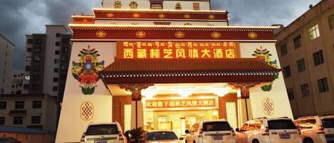 A hotel entrance with a traditional architectural design, featuring a prominent sign and a parking area with several cars.