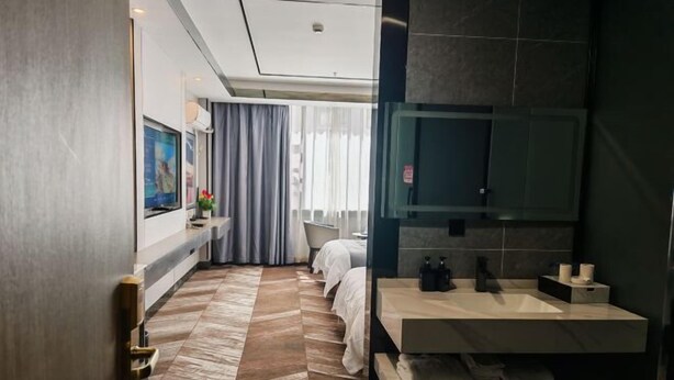 Moderni hotellihuone, jossa on suuri sänky, oleskelualue tuolilla ja pienellä pöydällä, kylpyhuone, jossa on suuri peili ja pesuallas, sekä näkymä kaupunkiin ikkunasta.