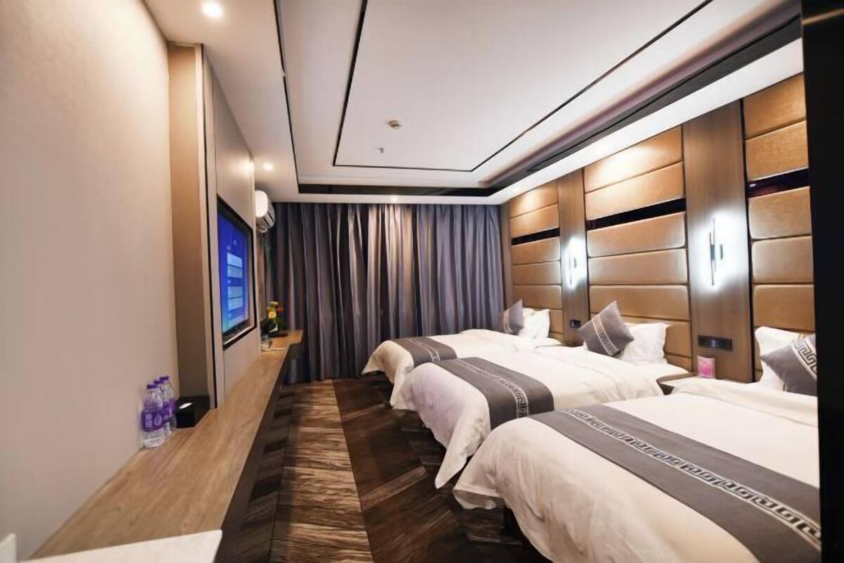 Quarto de hotel com três camas, televisão de tela plana, cabeceira de madeira e piso de madeira escura.