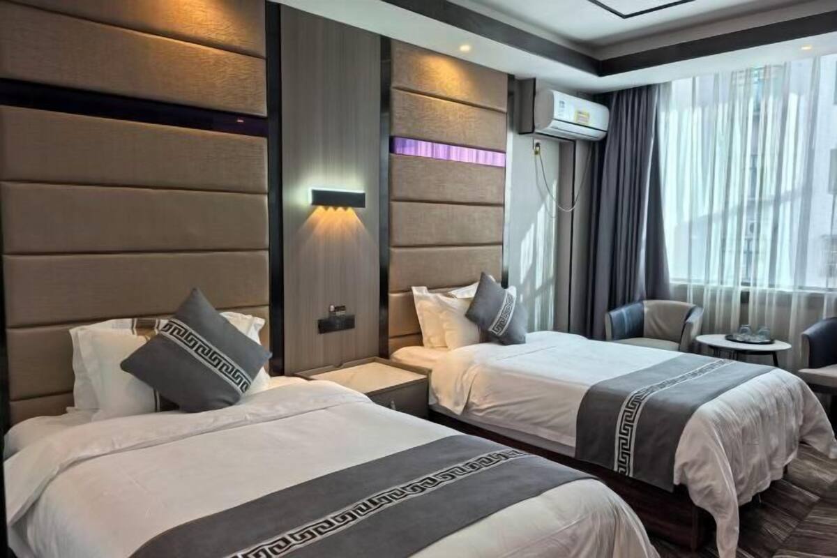 Quarto de hotel com duas camas, uma mesinha, uma cadeira e uma janela com cortinas.