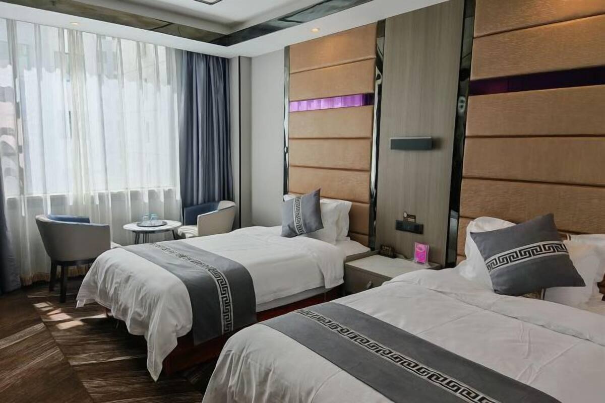 Quarto de hotel com duas camas, uma mesa pequena, uma cadeira e uma janela grande com cortinas.