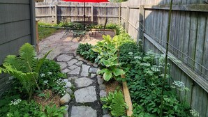 Un jardin avec un chemin de pierres, différentes plantes, une clôture en bois et un parasol rouge.