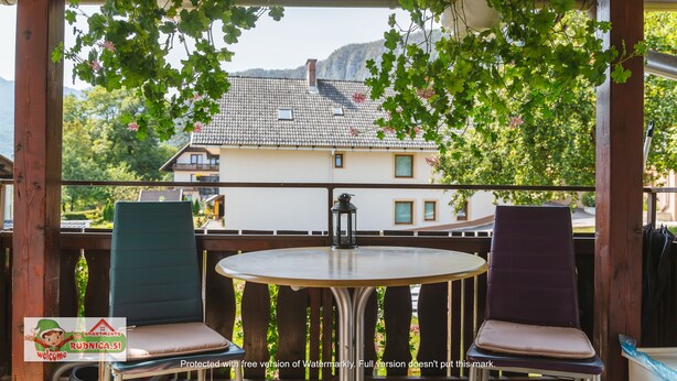 Eine Holzterrasse mit zwei Stühlen und einem Tisch, mit Blick auf ein Haus mit Ziegeldach und Grün im Hintergrund.