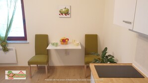 Ein kleiner, ordentlicher Raum mit zwei grünen Stühlen, einem weißen Tisch mit Obstschale und Tassen, einer Pflanze und einem Apfelbild an der Wand.