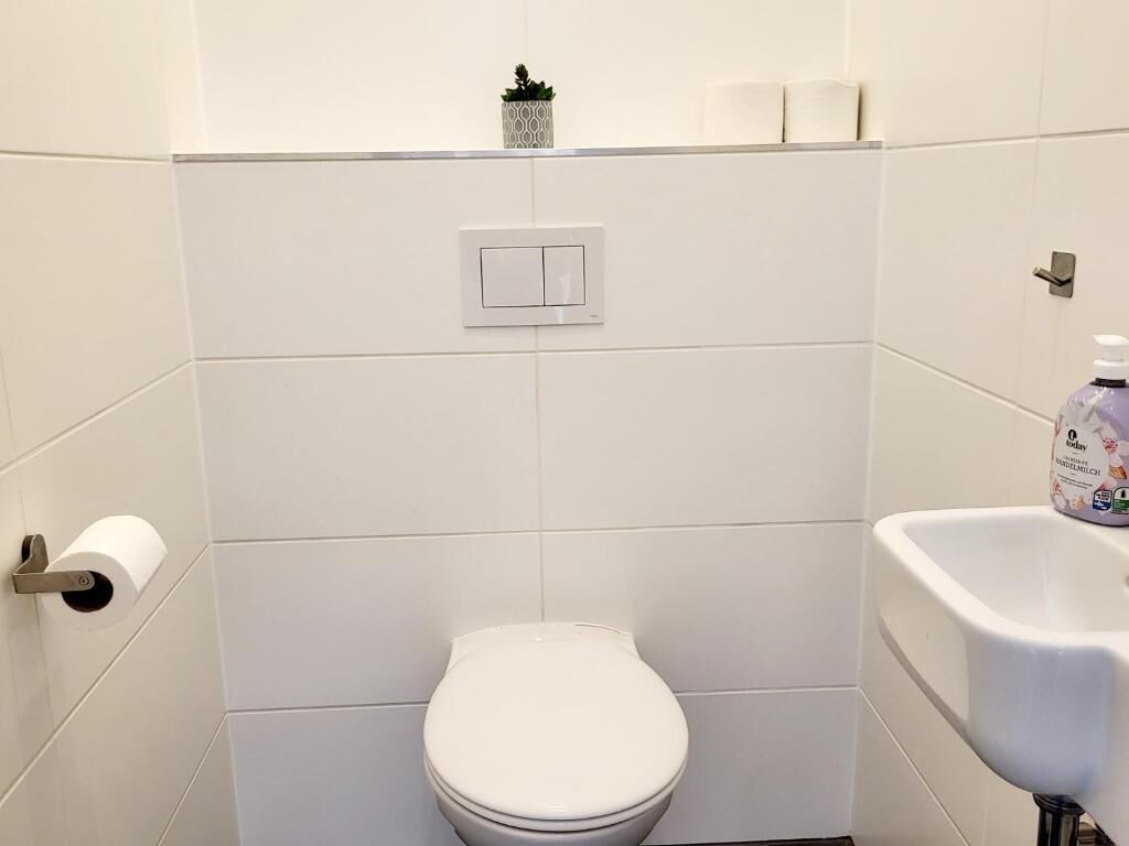 Une petite salle de bain aux murs recouverts de carreaux blancs, équipée de toilettes, d’un sèche-mains et d’un lavabo blanc.