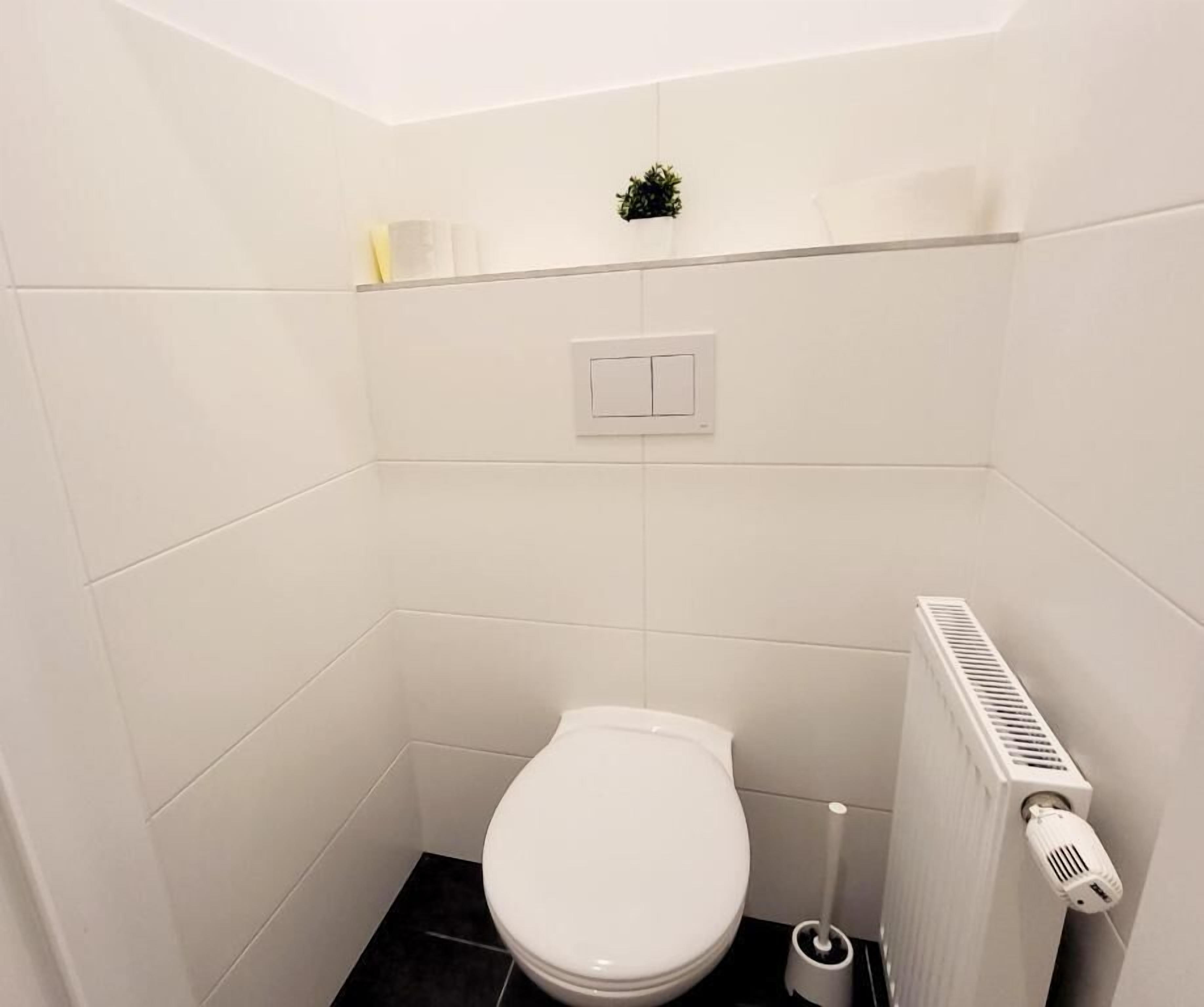 Une petite salle de bain aux murs blancs, avec des toilettes blanches, un radiateur et une petite plante accrochée au mur.