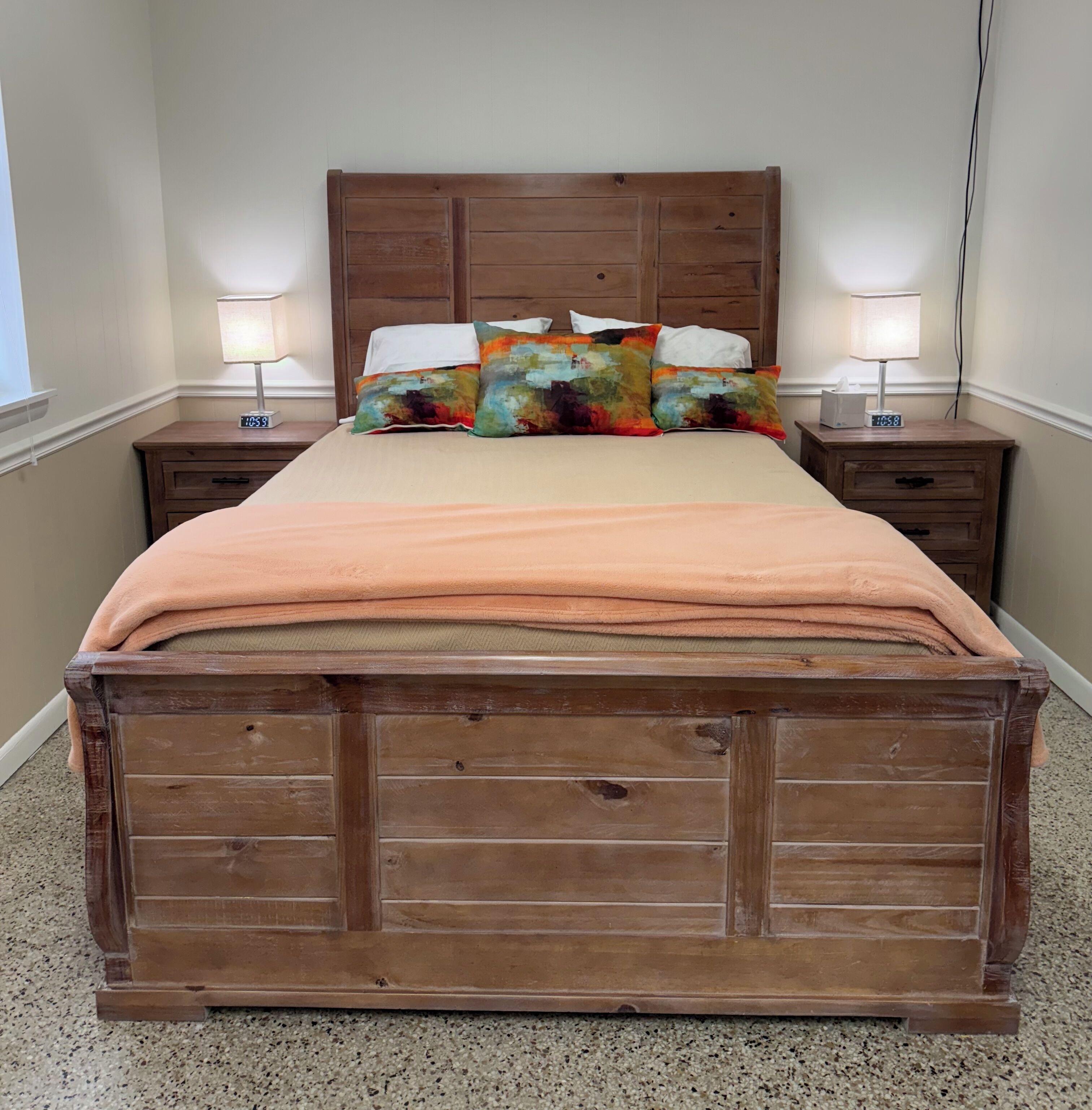 Una cama de madera con cabecera, dos mesitas de noche con lámparas y una ventana a la izquierda.