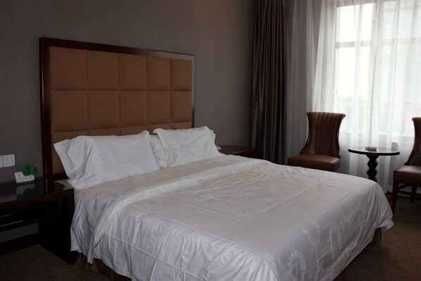 A hotel room with a large bed, a wooden headboard, a nightstand, a chair, a small table, and a window with curtains. - Wulate Hotel (Urad Front Banner)