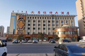 A hotel with a large sign, multiple floors, and a parking lot with cars. - Wulate Hotel (Urad Front Banner)